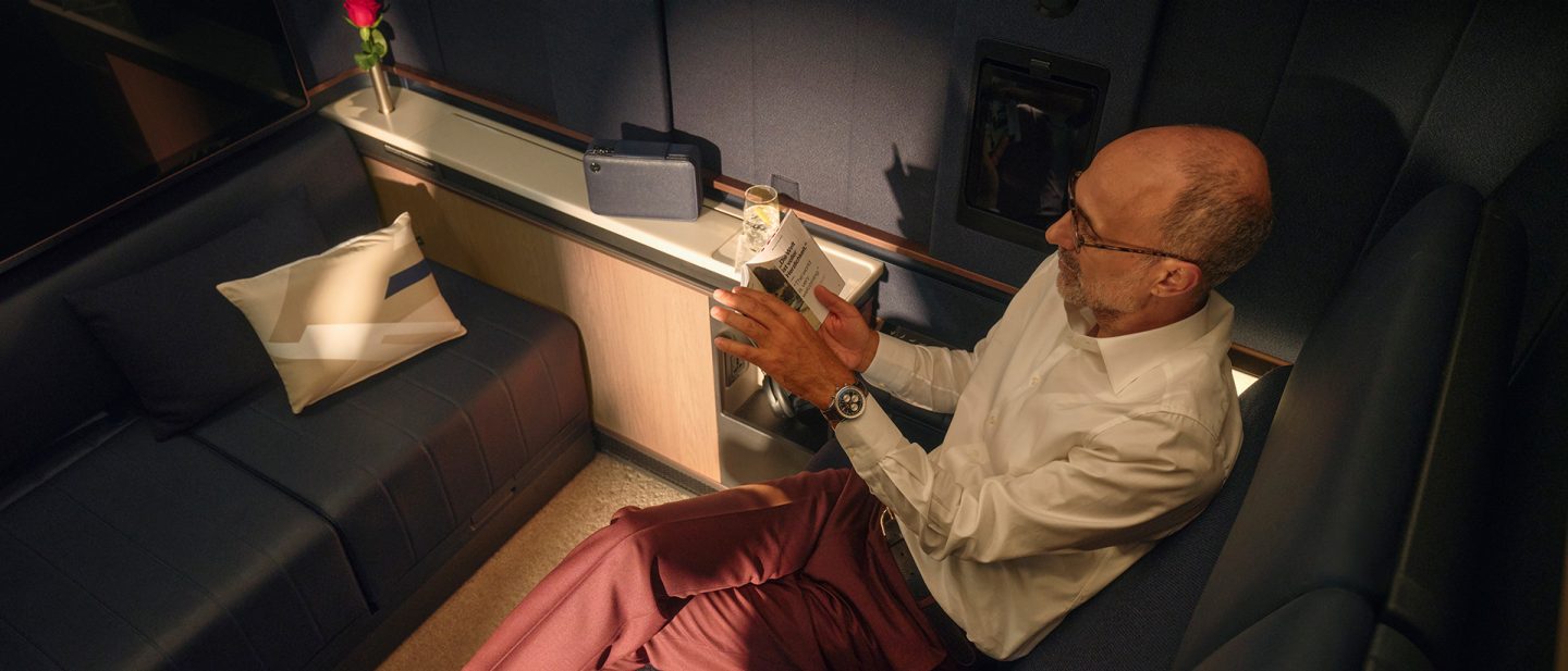 A man relaxes in a Lufthansa First Class suite, reading a menu of drinks, with a red rose and elegant cabin lighting