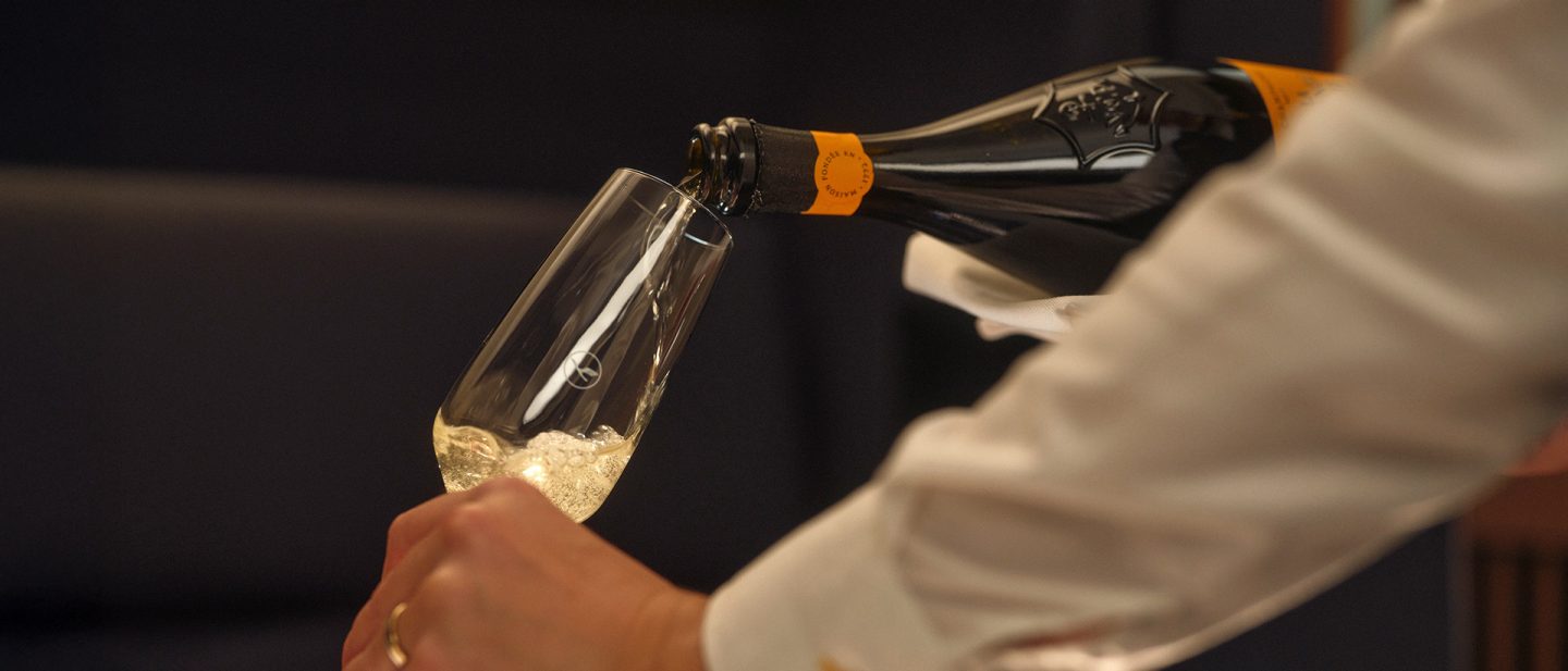 Flight attendant pouring champagne into a glass in a First Class cabin with soft ambient lighting