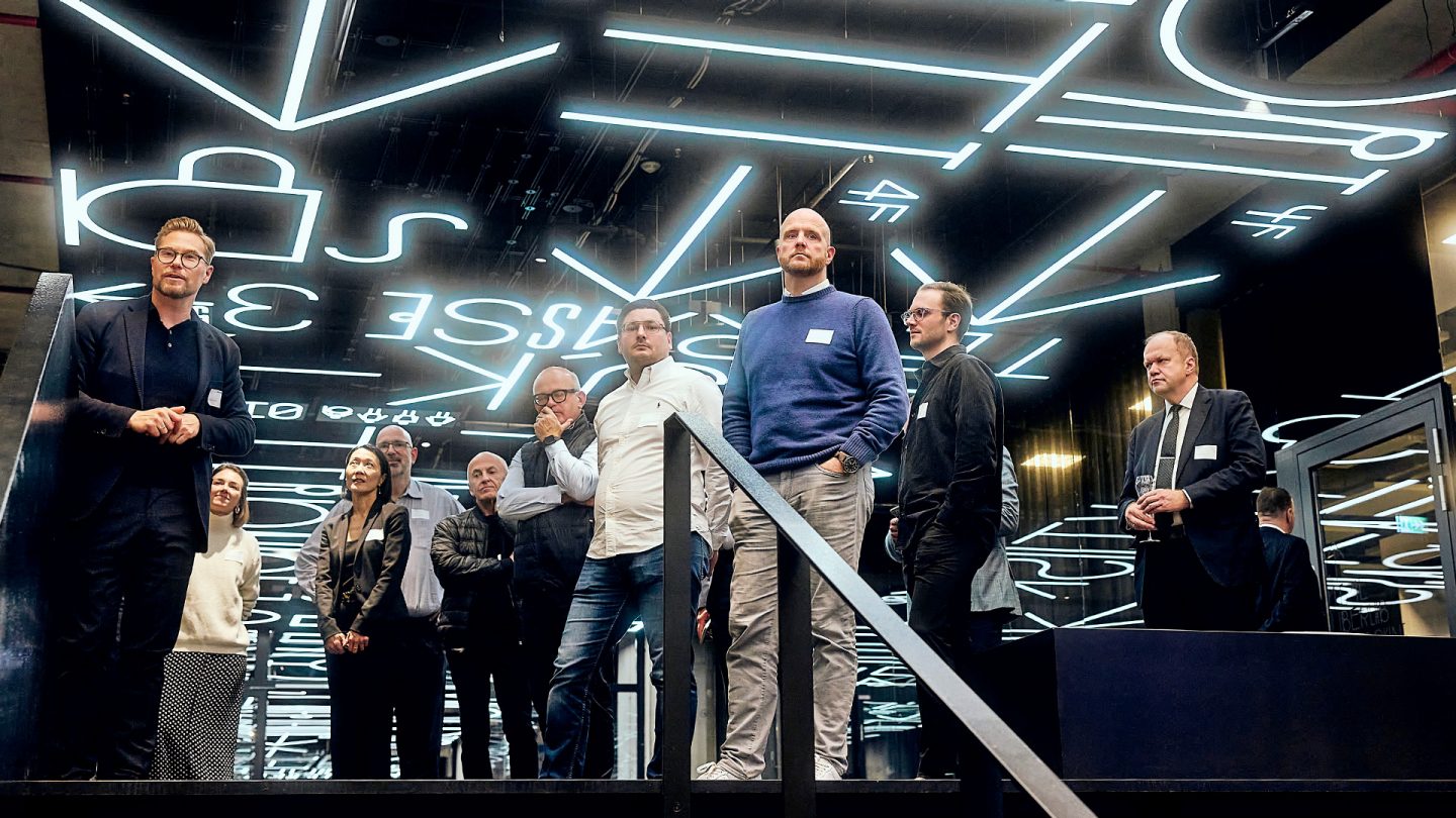 A group of people stands in a room with a futuristic, illuminated ceiling and listens attentively to a speaker.