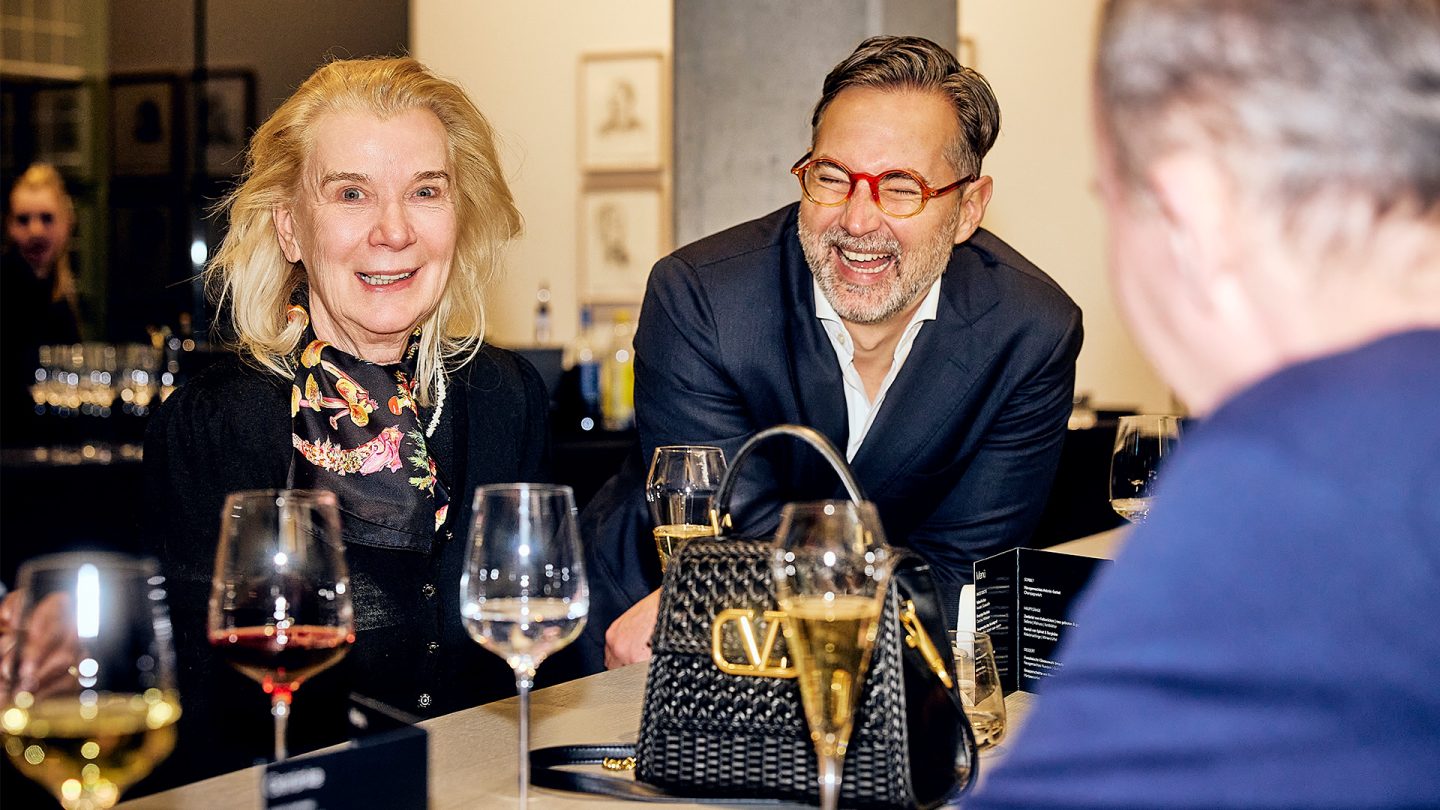 A woman and a man are sitting happily at a bar counter with several filled wine glasses and a black designer handbag in the foreground.