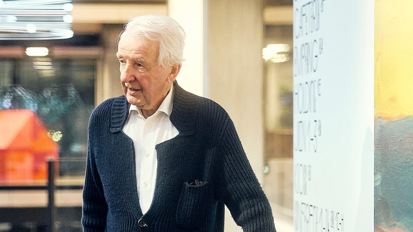An elderly man with white hair wearing a dark blue cardigan stands in front of a modern interior backdrop and looks at something outside the frame.