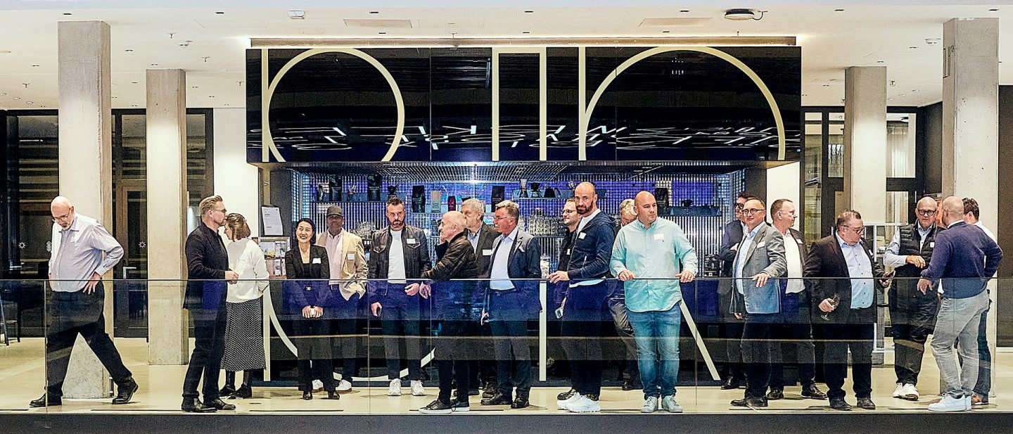 A group of elegantly dressed people stand in a modern building in front of a bar with a luminous ceiling installation.