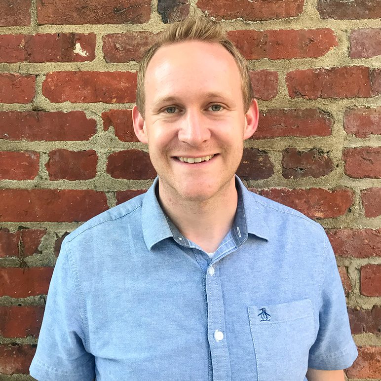 Photo of a smiling man in a blue shirt standing against a brick wall.