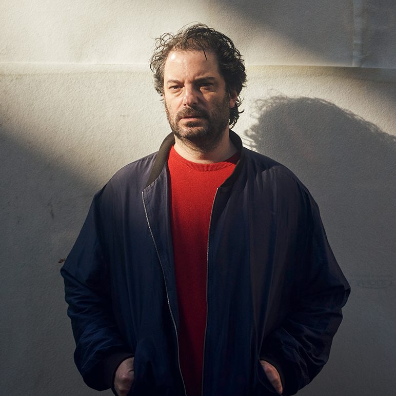 DJ Kalabrese in a red sweater and blue jacket standing against a sunlit wall, casting a shadow