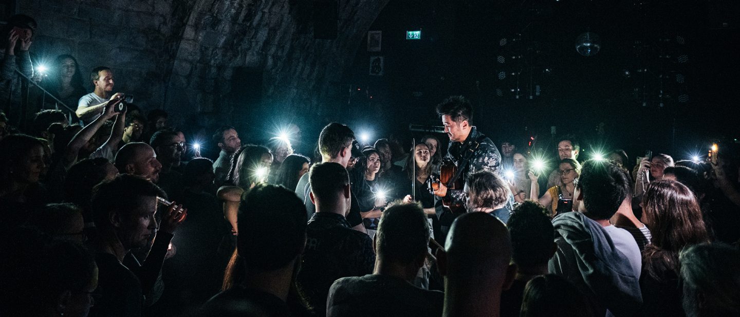 Live concert scene in the club Bogen F with a musician and audience members using smartphones as lights in a dark venue