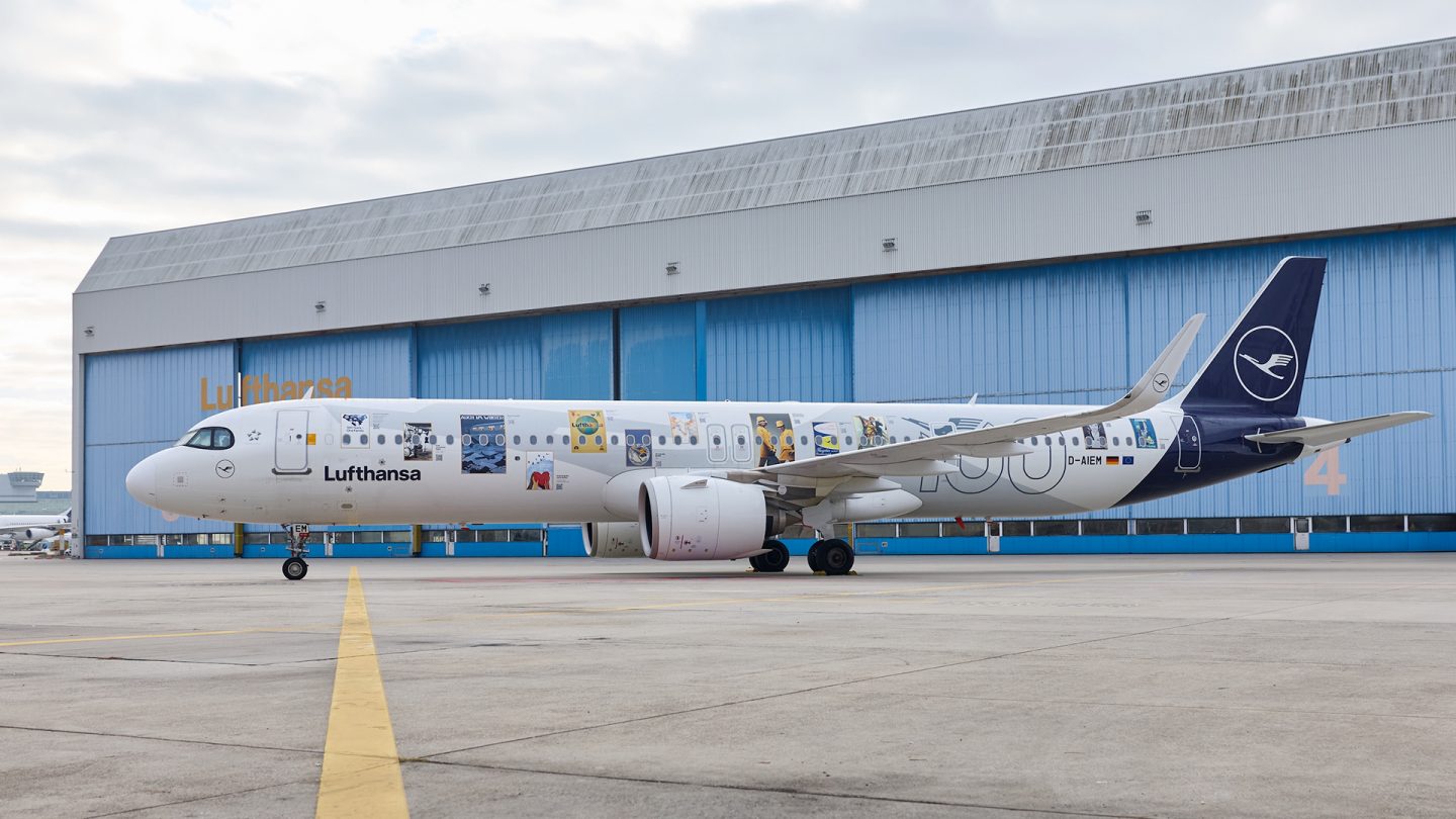 Technicians install retro posters on a Lufthansa aircraft fuselage in a maintenance hangar.