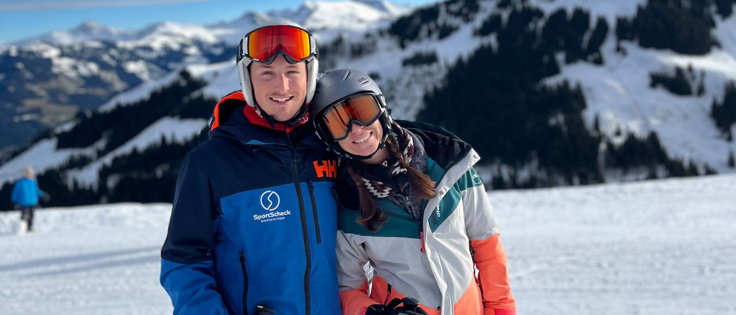 Couple in ski gear smiling close together in a snowy mountain landscape.