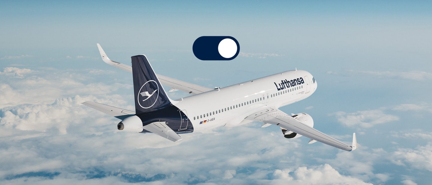 A Lufthansa commercial airliner flies above the clouds during the day, with a clear blue sky in the background.