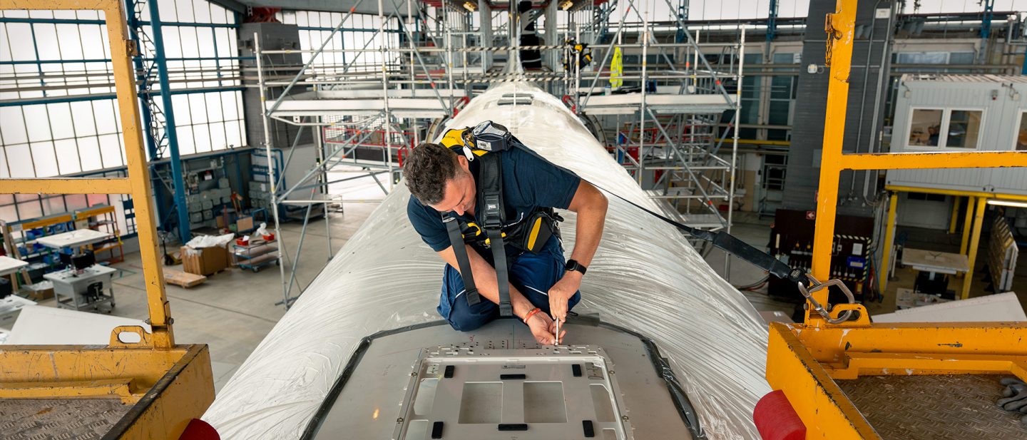 A technician wearing a safety harness is working on top of an airplane in a large hangar equipped with scaffolding.