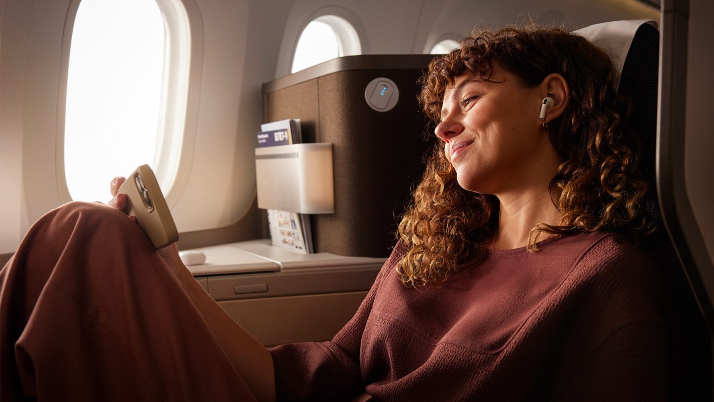 A woman with curly hair is listening to music with wireless earbuds while sitting in a business-class seat by the airplane window.