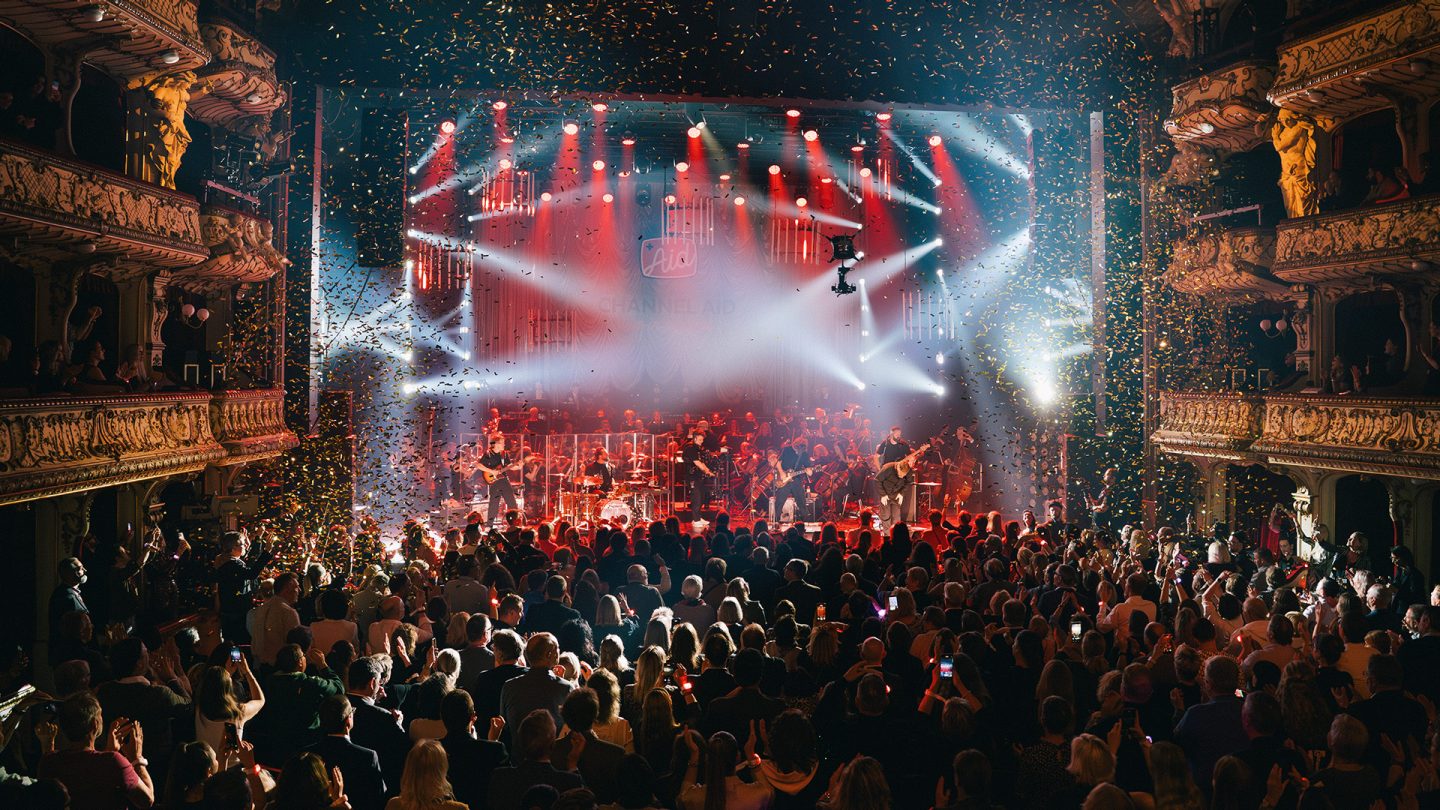 A concert in a glamorous theater hall with a band, orchestra, spotlights, and confetti