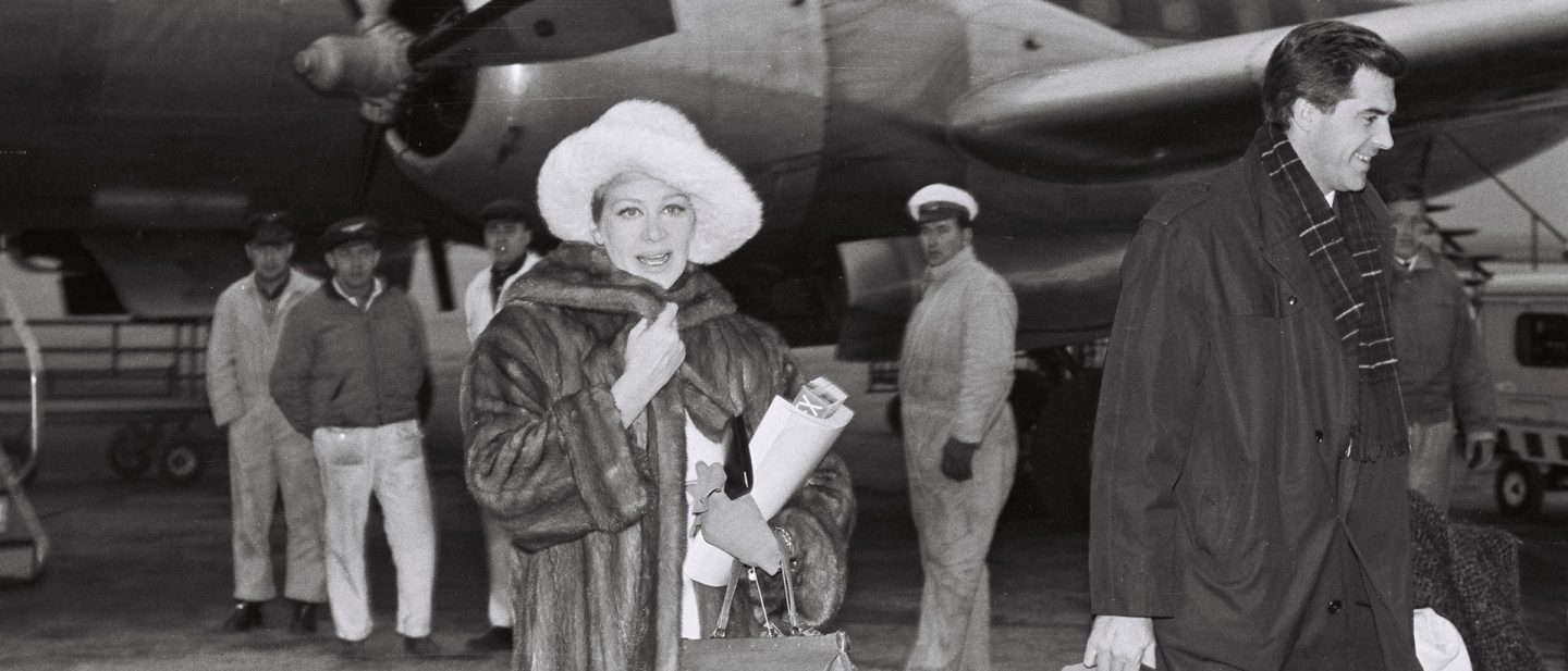 Historical black and white photo of an elegant woman in a fur coat and a man in front of a plane