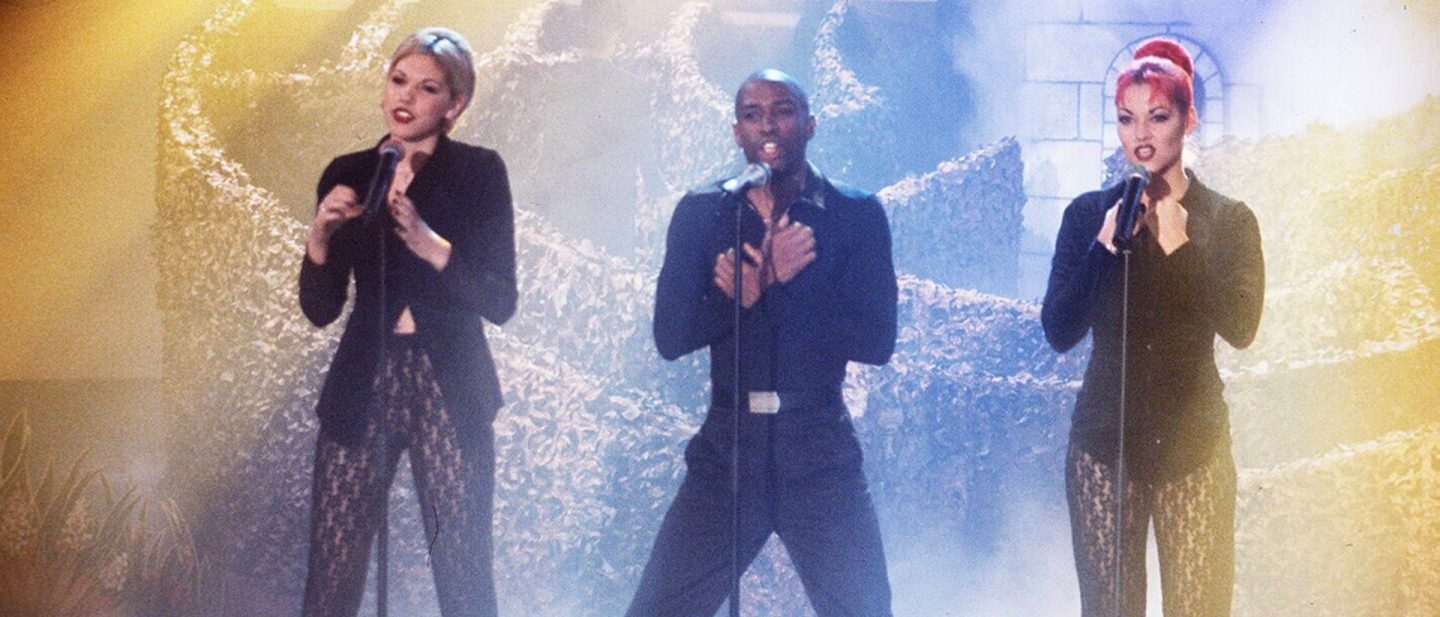 Three vocal artists in dark clothing performing live on a stage