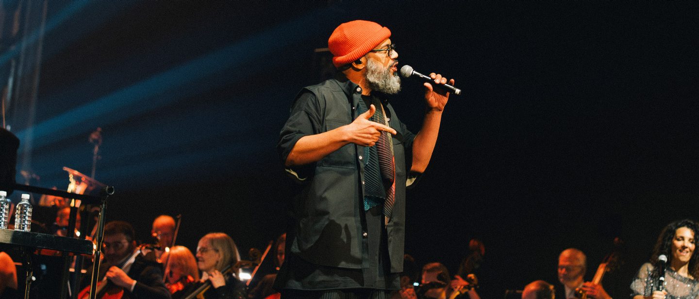 A singer with a red beanie and beard performs live on stage with an orchestra in the background