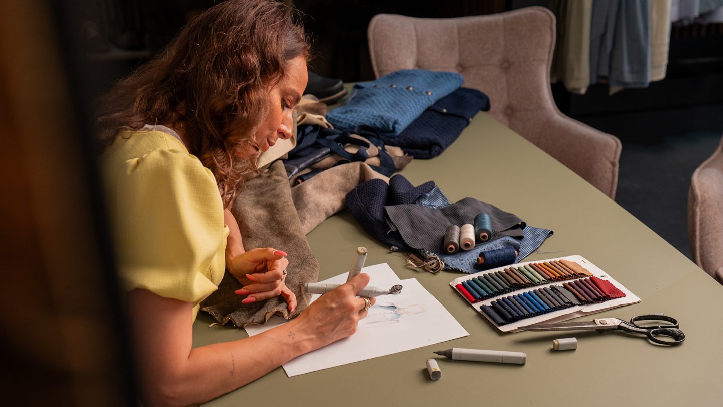 A fashion designer sketches at a table with fabrics, threads, and a color swatch card