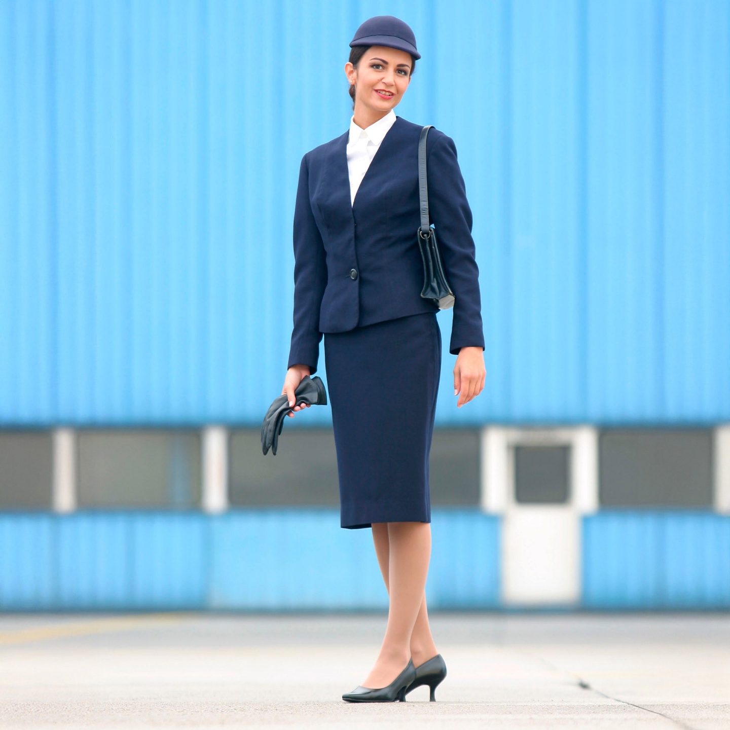 Flight attendant in navy Lufthansa uniform with hat, fitted skirt, and shoulder bag.