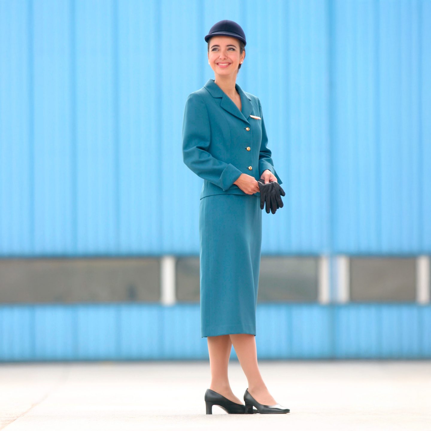 Flight attendant in turquoise Lufthansa uniform with hat, knee-length skirt, and black gloves.