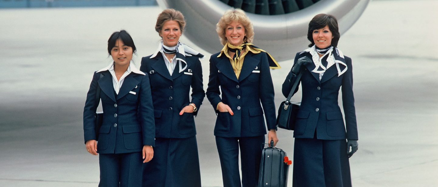 Four flight attendants in navy uniforms walk in front of an aircraft engine.