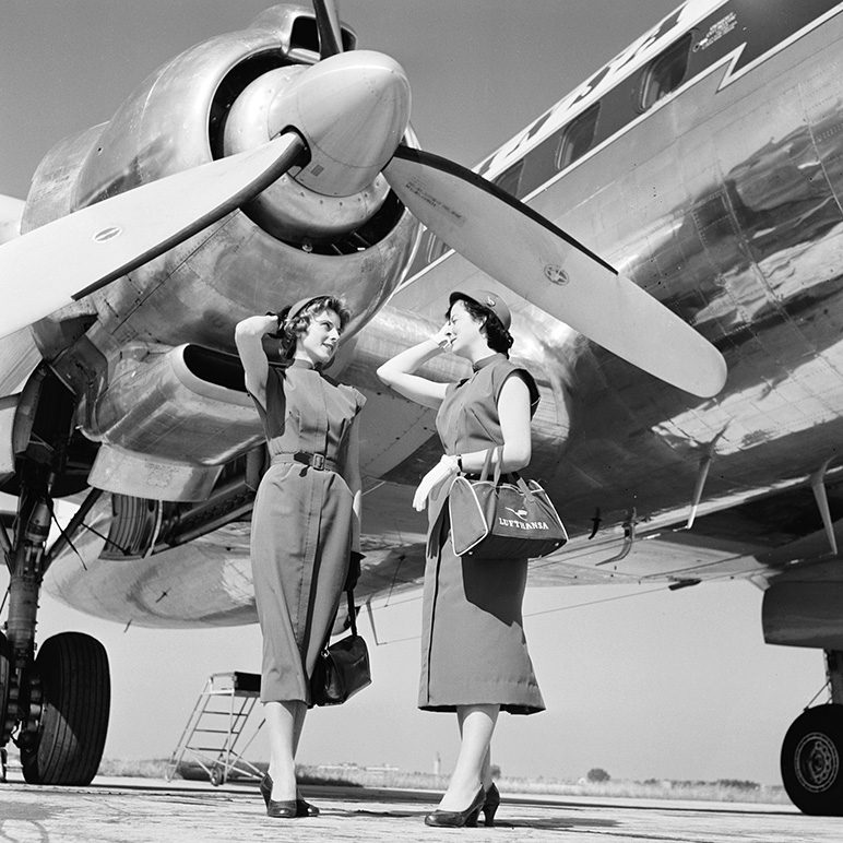 Two flight attendants pose beside a propeller aircraft in uniforms from the past.