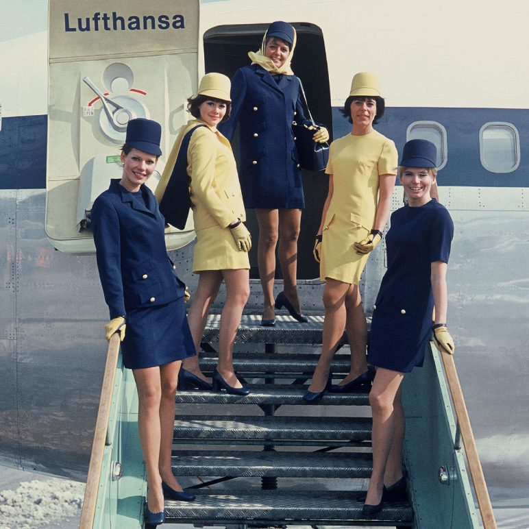 Lufthansa flight attendants in yellow and blue uniforms stand on aircraft stairs.
