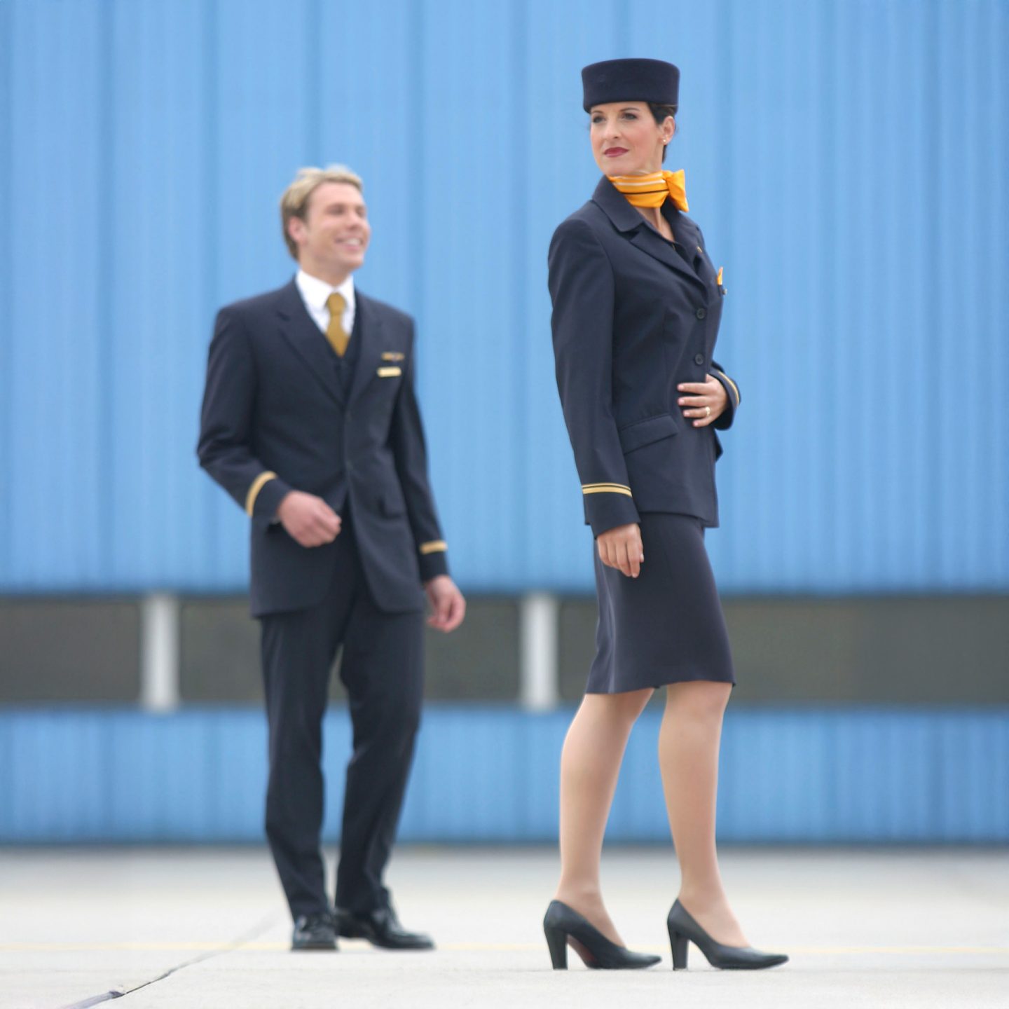 Flight attendant in navy Lufthansa uniform with yellow scarf and hat, male cabin crew in background.