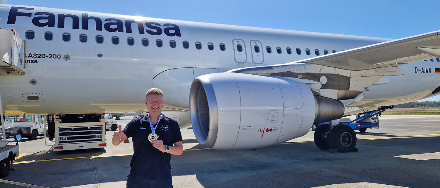 Man standing in front of an airplane with the inscription Fanhansa and holding a silver medal up to the camera