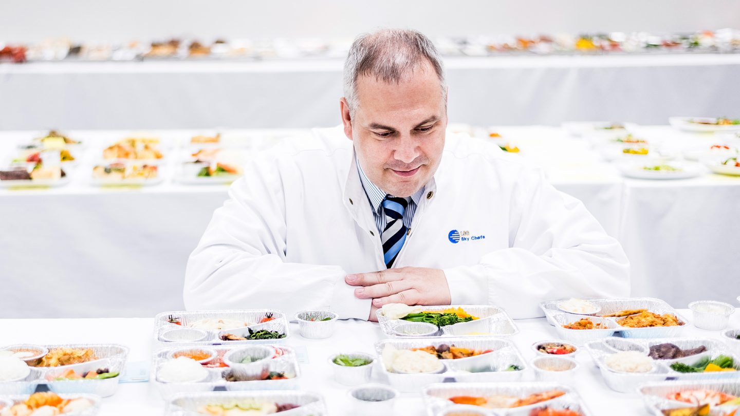 Man in a white coat sitting in front of many different airplane meals.