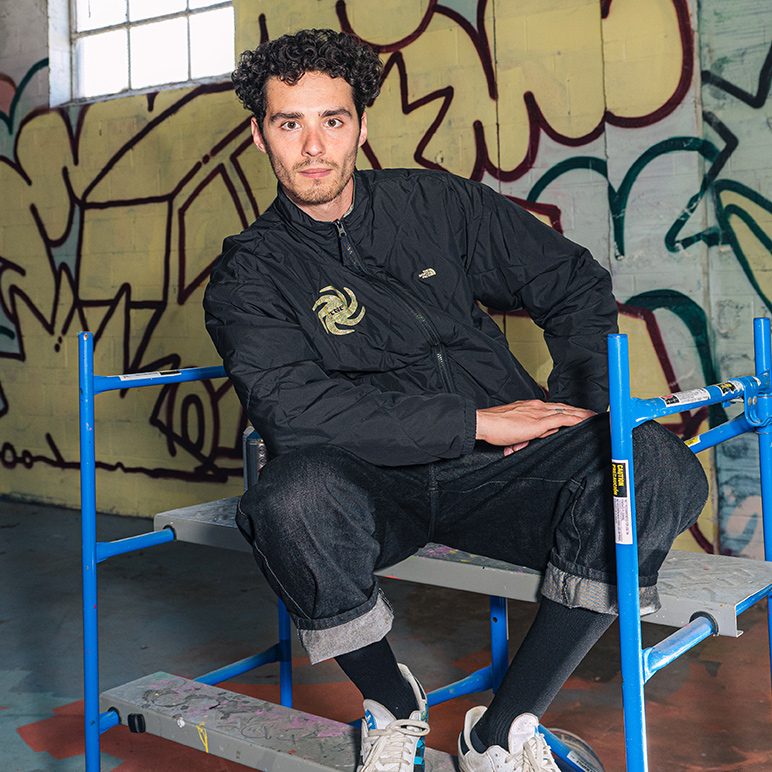 Artist in black clothing sits on scaffolding in front of graffiti-covered wall
