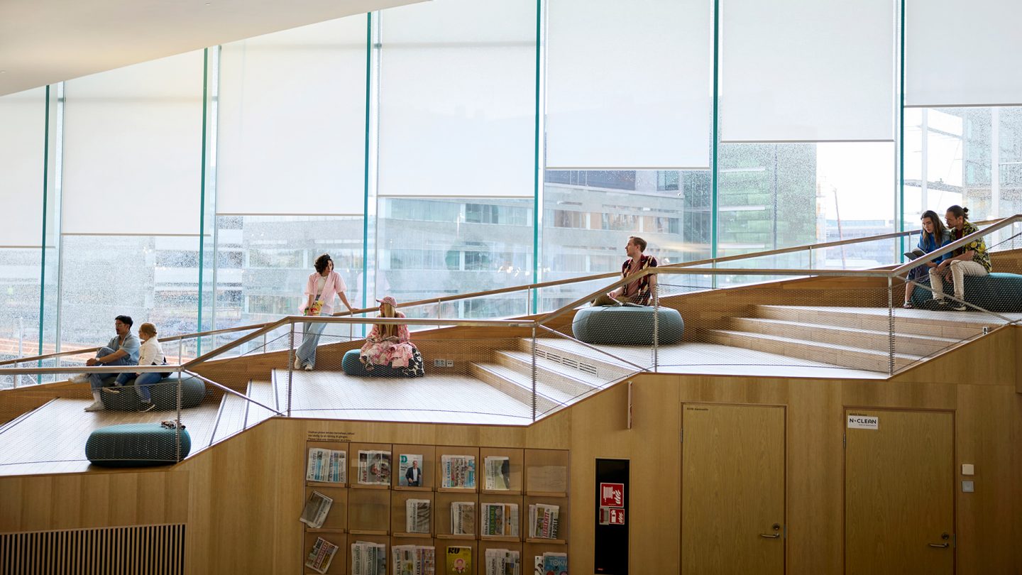 People relaxing and reading in a modern library with large windows and tiered seating areas.