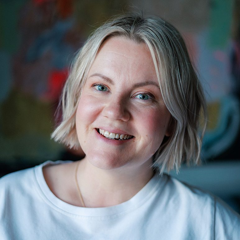 Portrait of a smiling woman with short blonde hair, wearing a white T-shirt, in front of a colorful background.