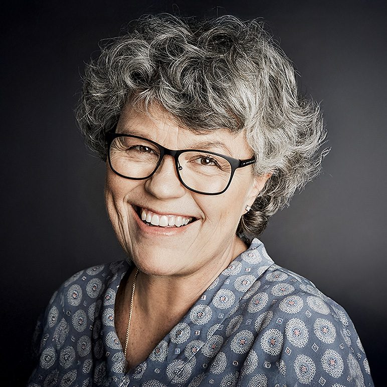 Studio portrait of Anne Holt with curly gray hair and black glasses against a dark background