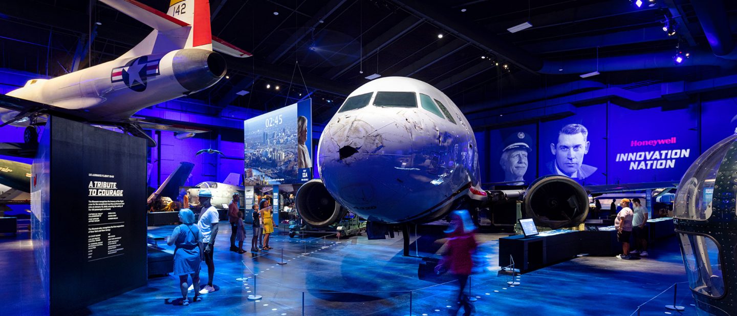 A blue-lit museum exhibit displays a large airplane nose with visible damage. Visitors observe, conveying curiosity.