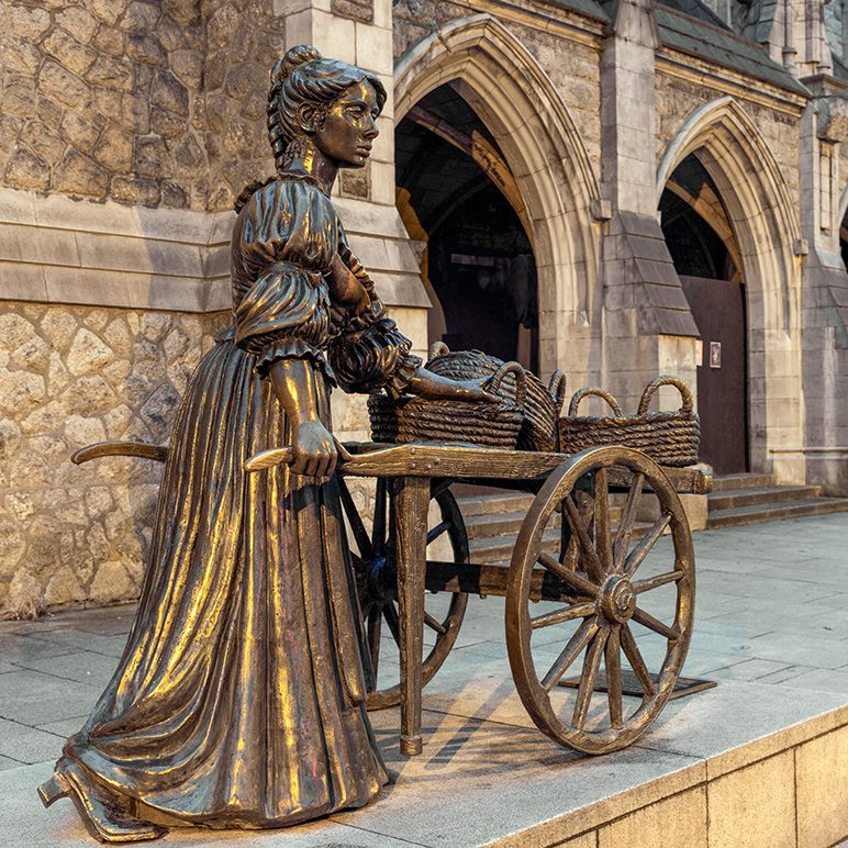 Bronze statue of a woman with a cart and baskets in front of a stone church in Dublin