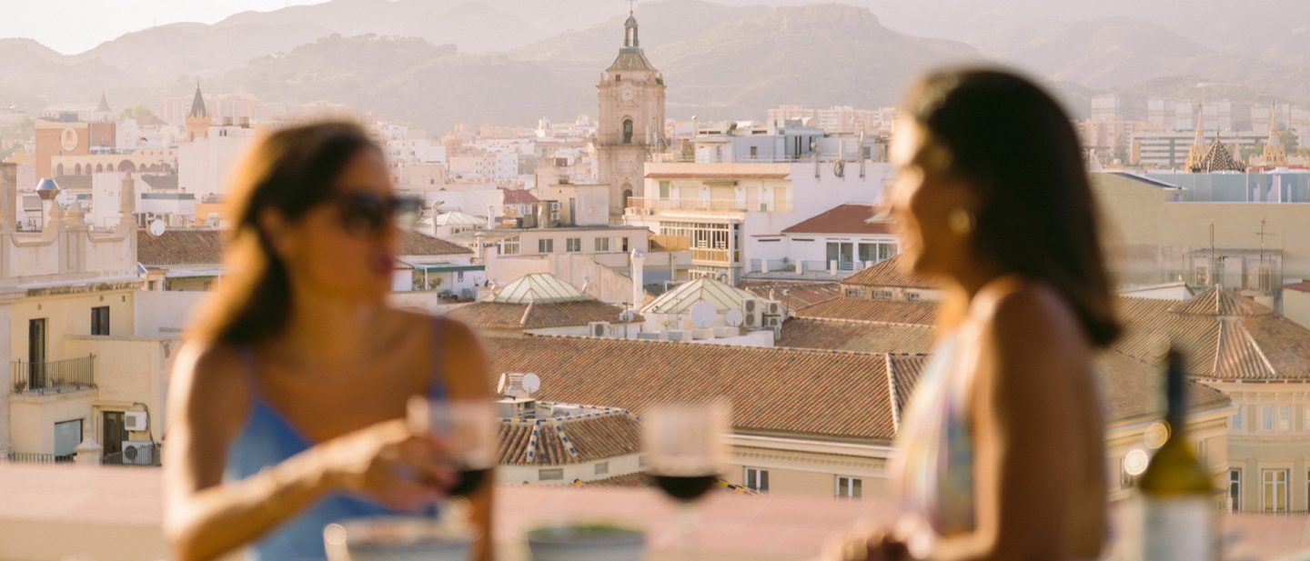 Zwei Frauen mit Gläsern auf einer Dachterrasse vor der Stadtkulisse von Málaga