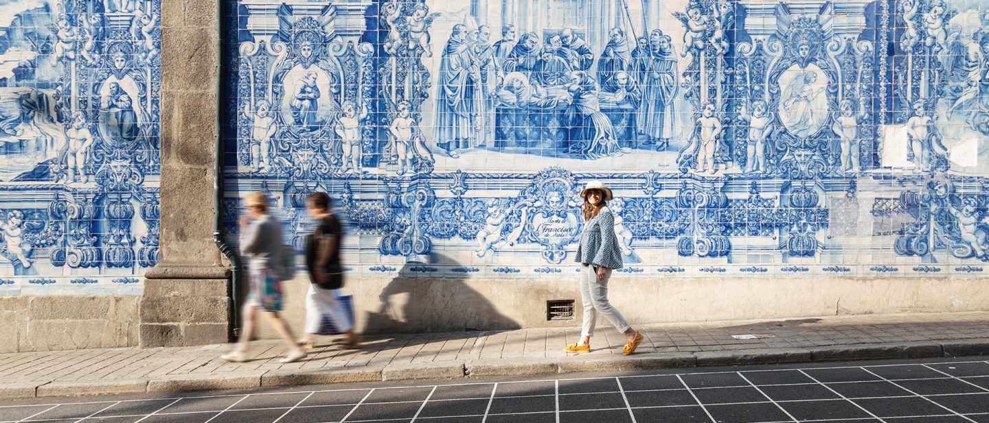 Woman in a sun hat walking past a large blue and white azulejo tile wall in Portugal