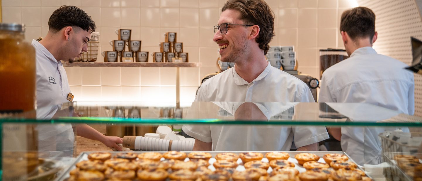 Three bakers in white uniforms behind a counter with fresh pastéis de nata