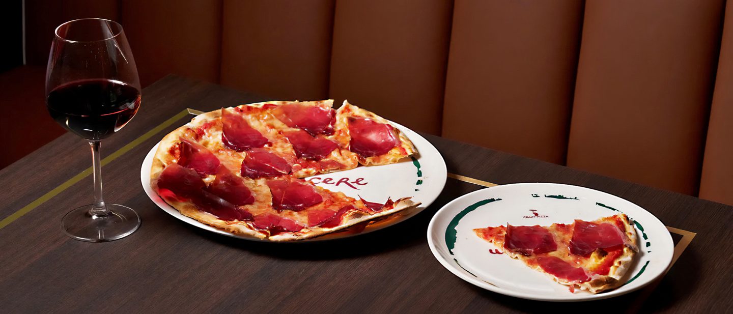 Pizza with prosciutto next to a glass of red wine on a dark wooden table