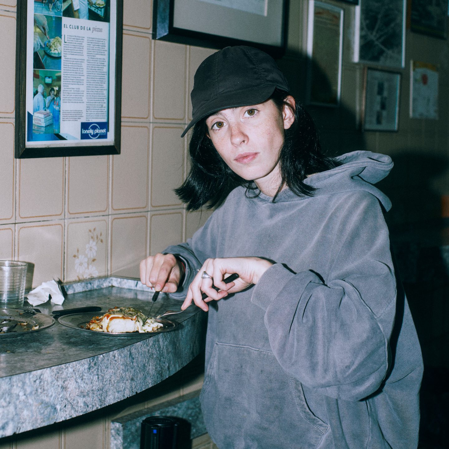 Franzizca is standing at a bar table wearing a gray hoodie and eating pizza.