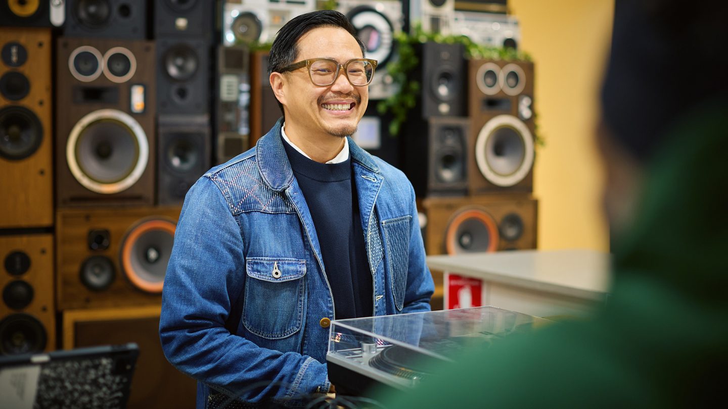 Oliver Mak, der eine Brille und eine Jeansjacke trägt, steht vor einer Wand aus Lautsprechern und lächelt
