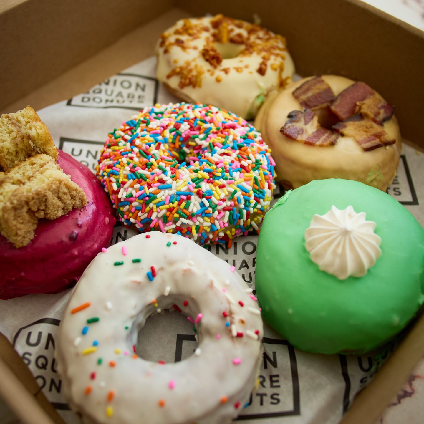 Eine Schachtel mit sechs Donuts, darunter Sorten mit bunten Streuseln, Speck, grünem Guss und Beeren