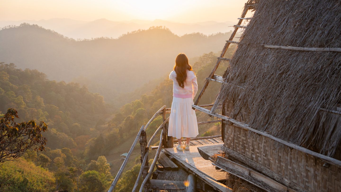 A woman looks out from a wooden hut at forested mountains in the golden morning light