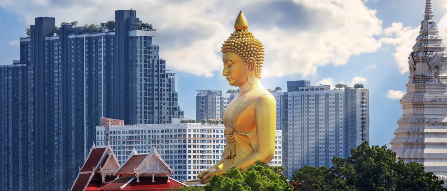Giant golden Buddha statue overlooking modern cityscape with skyscrapers and traditional buildings.