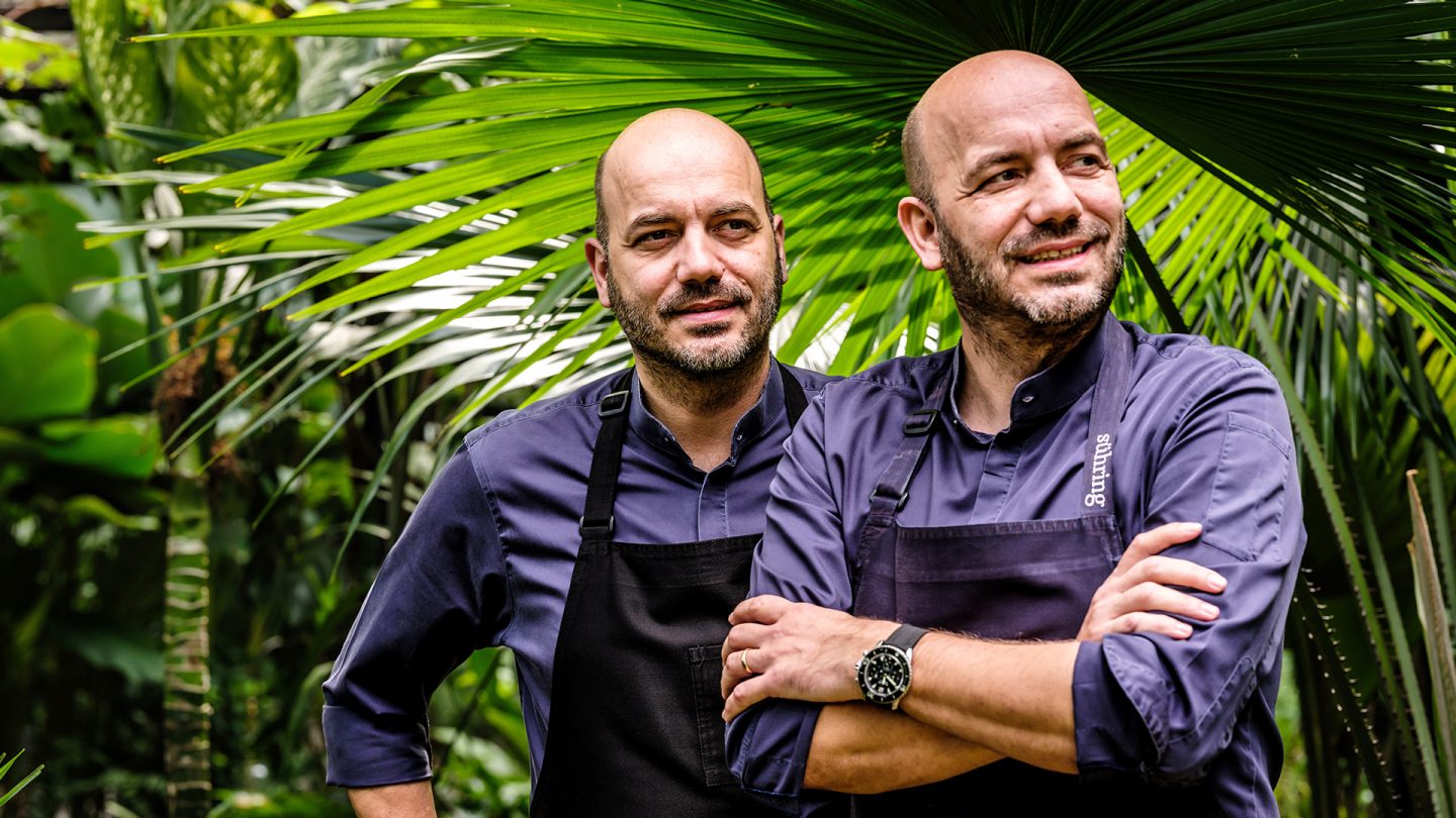 Two male chefs in aprons standing with arms crossed in a lush green garden.