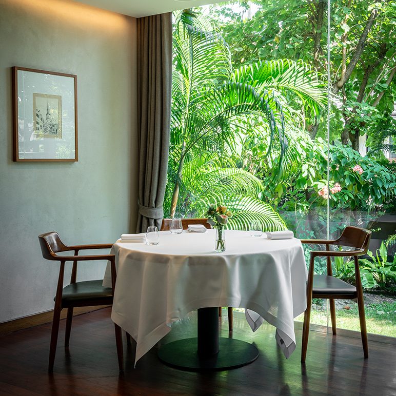 Elegant dining area with large windows overlooking a lush garden, modern wooden chairs, and a white tablecloth.