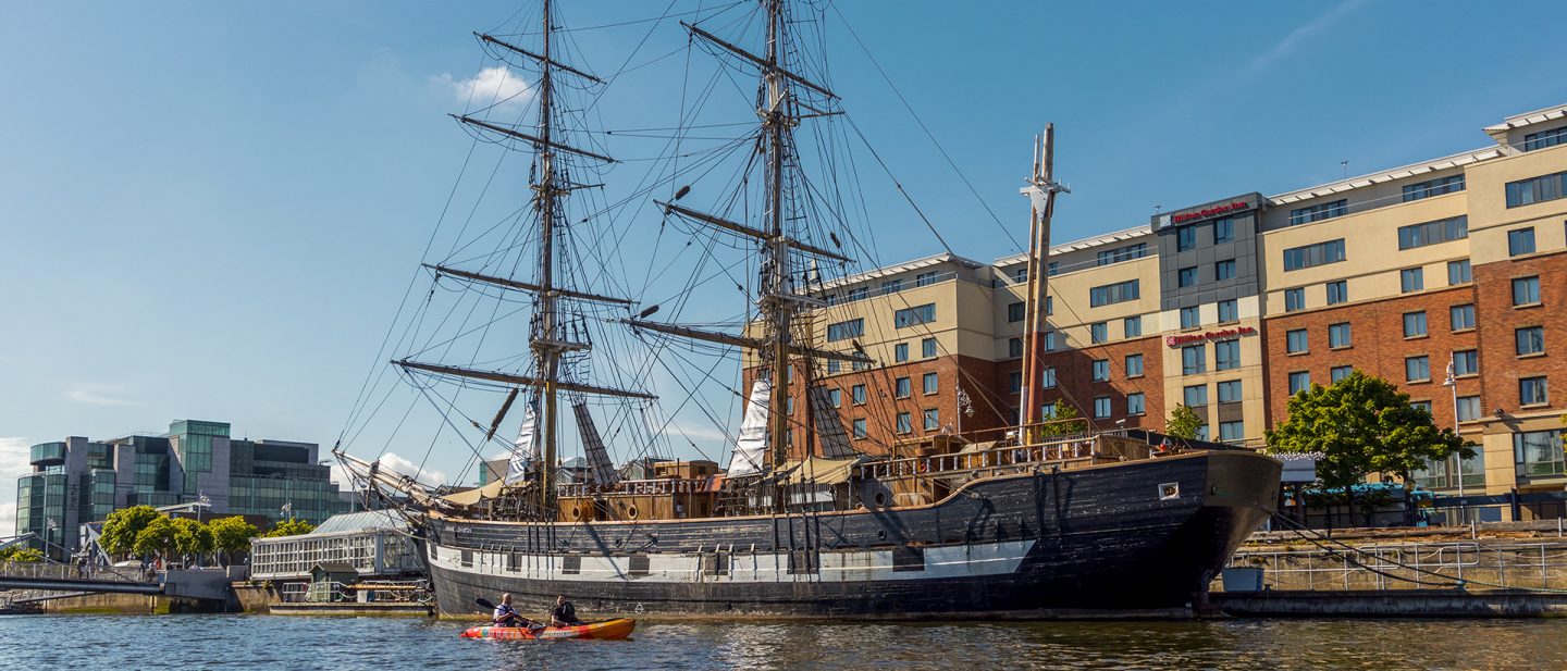 Tall ship docked beside modern buildings, with kayakers nearby, on a sunny day in Dublin