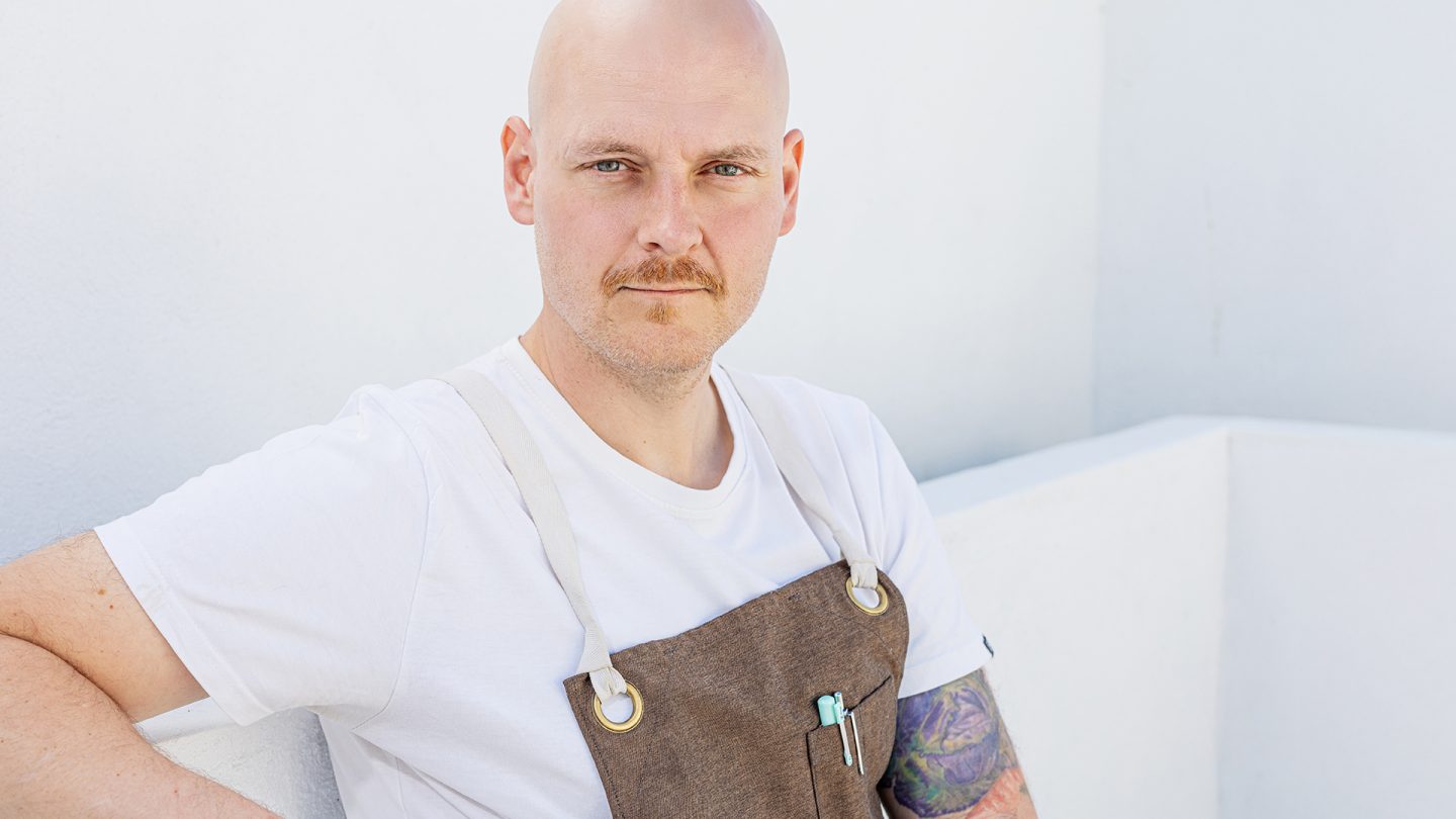 Malte Half, wearing a white shirt and a brown apron, leans against a white wall