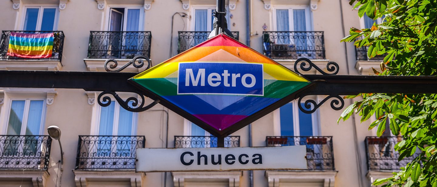 Metro Chueca station sign decorated with rainbow colors and an LGBTQ+ pride flag on a balcony in the background.