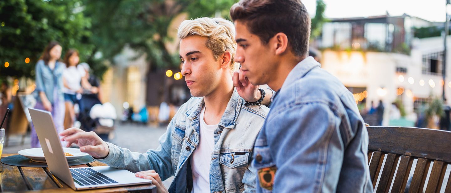 Two young men in denim jackets working on a laptop outdoors at a wooden table in an urban setting.
