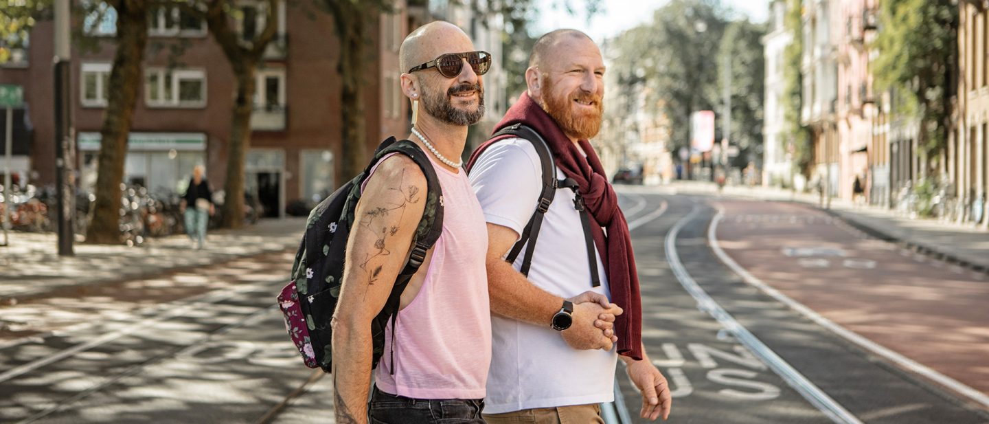 Two men with backpacks smiling and walking along a sunny city street with tram tracks in the background.