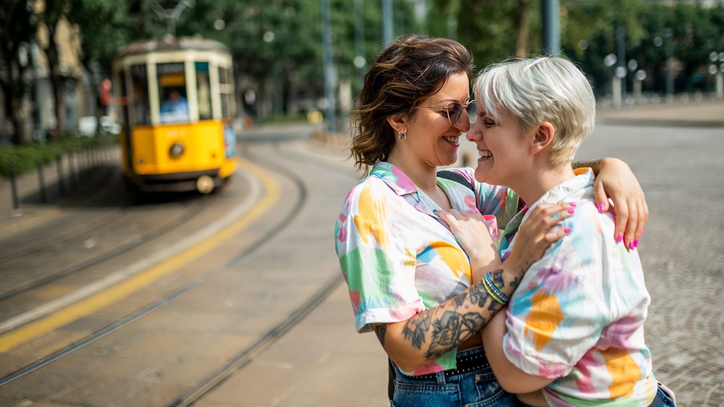 Two women wearing colorful shirts embrace and smile near tram tracks with a yellow tram in the background.